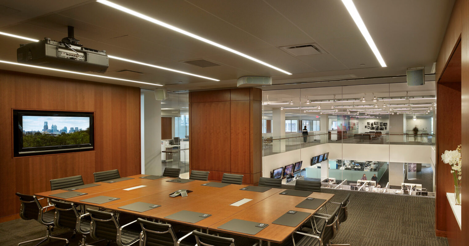 Modern conference room featuring sleek, rectangular table with built-in technology ports, surrounded by ergonomic chairs. Wood-paneled walls complement overhead linear LED lighting, with an adjacent open-office area visible through the glass partition, enhancing the sense of space.