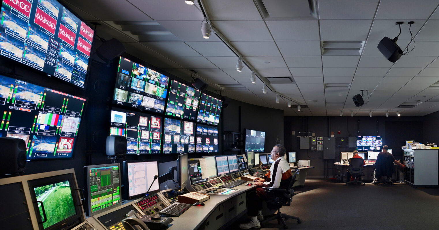 Modern broadcast control room with ergonomic design featuring multiple illuminated screens, technical consoles, and one operator in a dimly lit environment optimizing focus on digital content.