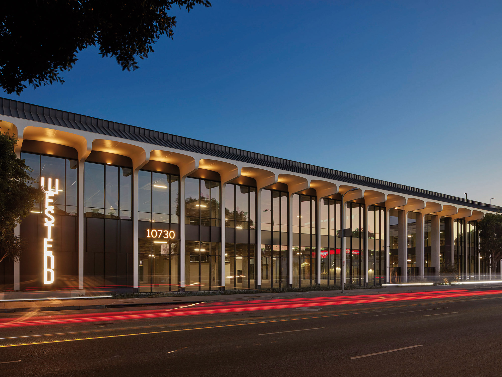 Modern commercial building with elegant columns and large windows illuminated at twilight, with the urban life's streaks of light from passing cars on the bustling street.