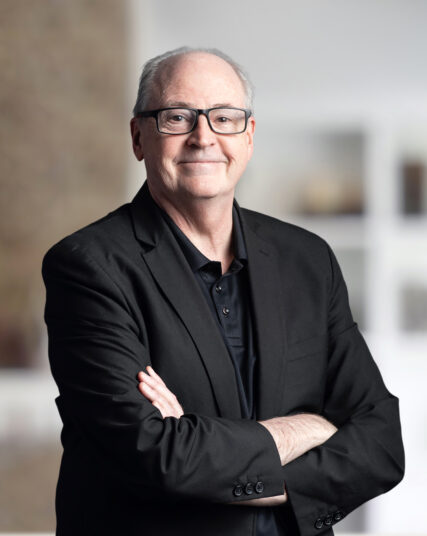 The image features a man with glasses and grey hair, dressed in a black shirt and blazer, with his arms crossed in a confident stance. The soft, neutral background suggests an indoor setting with a professional atmosphere.