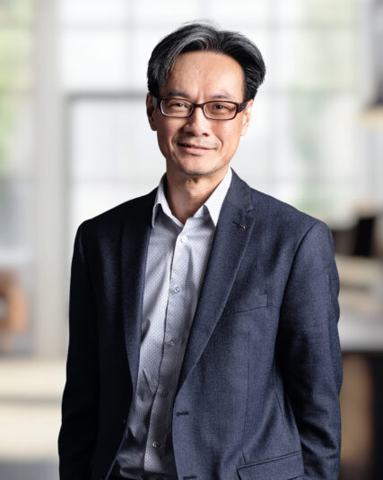 The image captures a man with glasses and neatly combed hair, offering a gentle smile, dressed in a tailored dark blazer over a light patterned shirt. He stands against a backdrop of light diffused through windows, suggesting a bright and contemporary office environment.