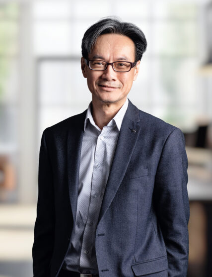 The image captures a man with glasses and neatly combed hair, offering a gentle smile, dressed in a tailored dark blazer over a light patterned shirt. He stands against a backdrop of light diffused through windows, suggesting a bright and contemporary office environment.