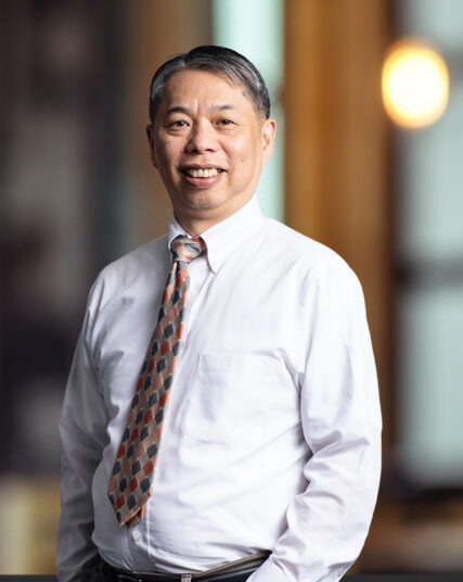 The photo captures a man with a friendly demeanor, wearing a crisp white shirt, a checkered tie in shades of orange and grey, and a belt. He stands with his hands casually in his pockets, against an out-of-focus background illuminated by warm light, indicative of an interior office space.