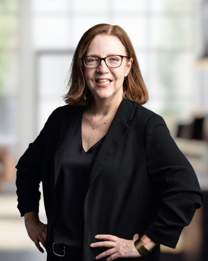 The photograph features a woman with medium-length hair and glasses, smiling in a black ensemble that includes a v-neck top and a blazer. Her hands rest lightly on her hips as she stands in an office space, with light streaming in from the background, illuminating her with a soft glow.