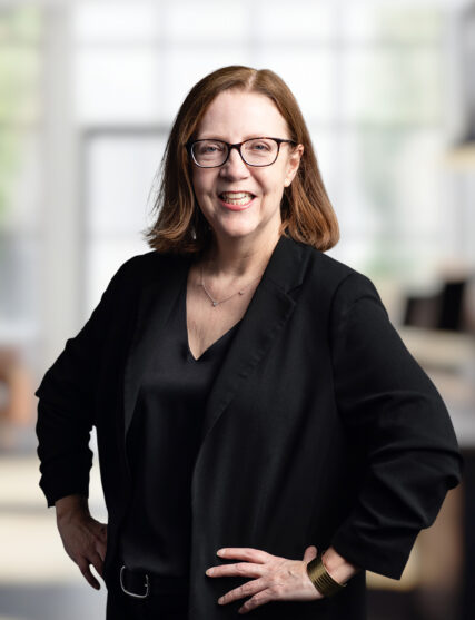The photograph features a woman with medium-length hair and glasses, smiling in a black ensemble that includes a v-neck top and a blazer. Her hands rest lightly on her hips as she stands in an office space, with light streaming in from the background, illuminating her with a soft glow.