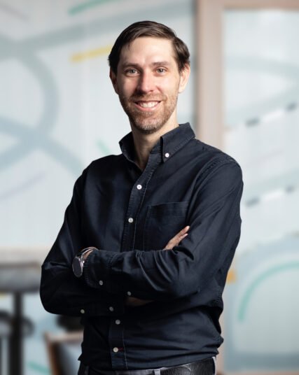 The image features a man with a slender build and a warm, engaging smile, dressed in a casual, dark button-down shirt with his arms confidently crossed. The background is light and abstract, suggestive of a creative or design-focused workplace environment.