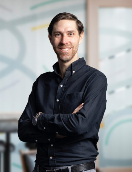 The image features a man with a slender build and a warm, engaging smile, dressed in a casual, dark button-down shirt with his arms confidently crossed. The background is light and abstract, suggestive of a creative or design-focused workplace environment.