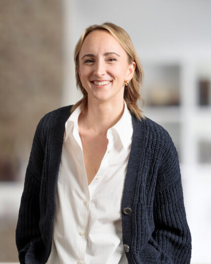 The image shows a woman with shoulder-length blonde hair and a warm smile, wearing a white blouse and a navy blue cardigan. The soft-focus background suggests a bright and spacious interior, possibly an office or gallery space.