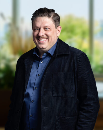 The image displays a man with a cheerful expression and neatly styled hair, dressed in a blue shirt and a dark textured coat. He stands with a slight lean forward, against a backdrop of blurred natural elements and daylight, giving the impression of a comfortable and inviting workspace.