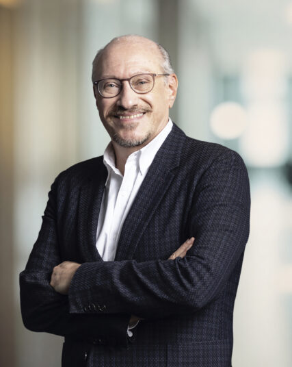 The image shows a man with a beard and glasses, sporting a confident smile, dressed in a dark textured blazer over a crisp white shirt. He stands with his arms folded, projecting a relaxed yet authoritative demeanor, against a softly blurred background typical of a professional office environment.