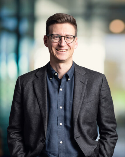 The image features a smiling man with glasses, short hair, and a smart-casual appearance, wearing a dark grey blazer over a navy shirt. The background has a bokeh effect with soft, circular light spots, suggesting a modern and dynamic office environment.