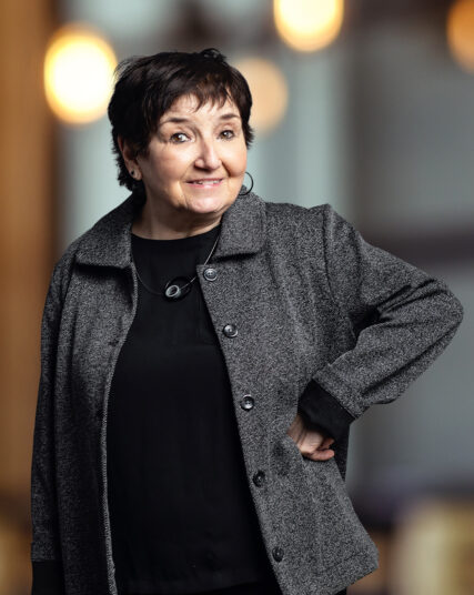 The photo depicts a woman with a playful expression and short, dark hair, dressed in a black outfit with a textured grey coat. She is posed with one hand on her hip, standing against a backdrop that features warm, glowing bokeh lights, suggesting an elegant indoor atmosphere.