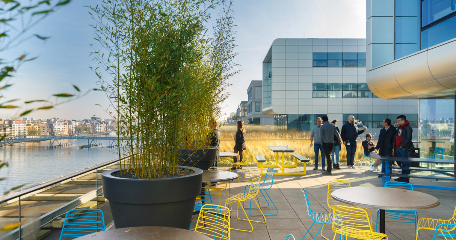 Modern terrace with vibrant yellow and lime green metal chairs, round tables, and large planters. Sleek glass building facade reflects the waterside setting, while visitors engage in conversation amidst the urban landscape.