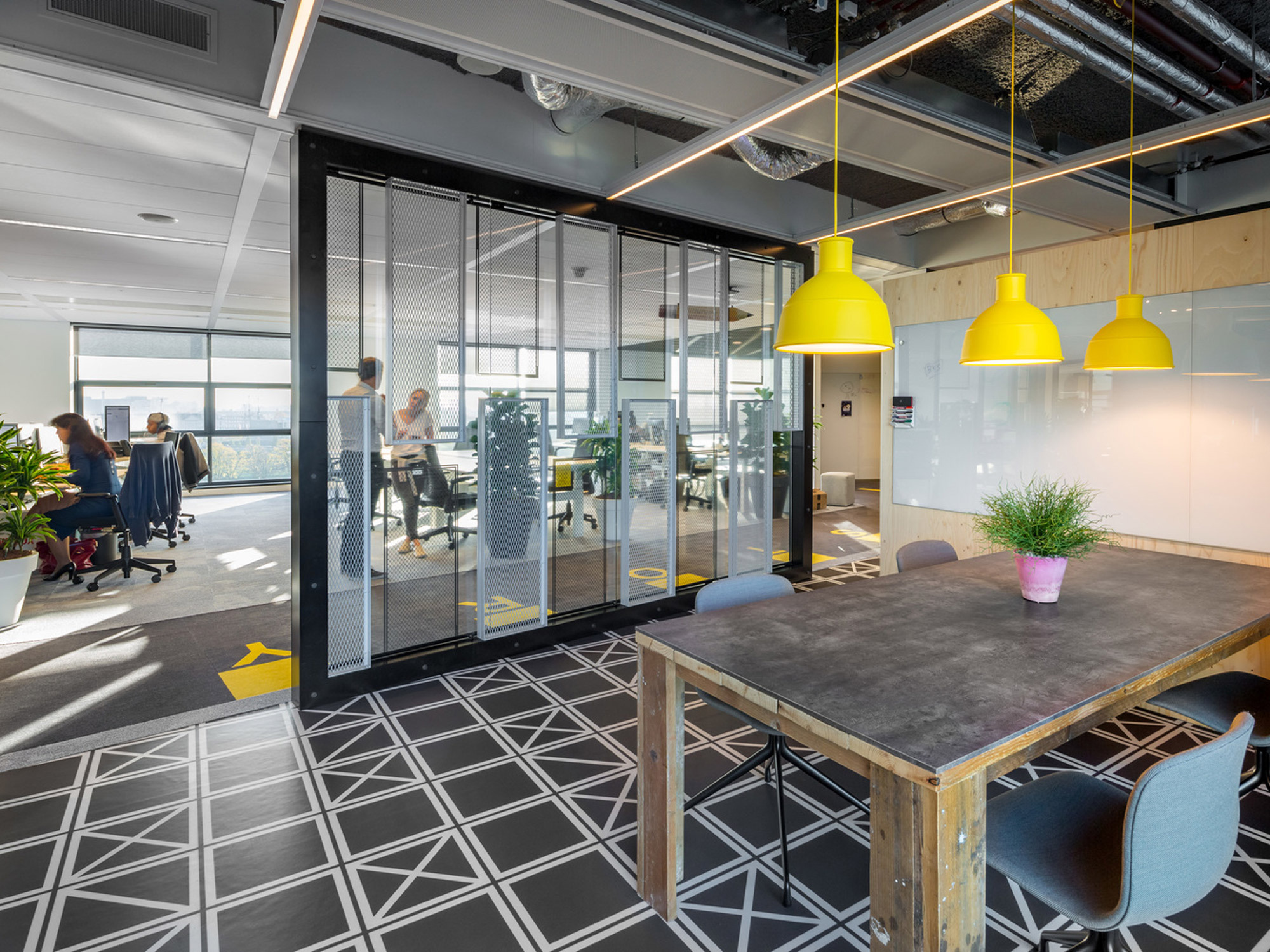 Modern office space with expansive views, featuring a geometric floor pattern, overhead yellow pendant lighting, and a glass divider framing a sleek, communal workspace. Natural light floods the area, enhancing the open-concept design and the collaborative environment punctuated by pops of greenery.