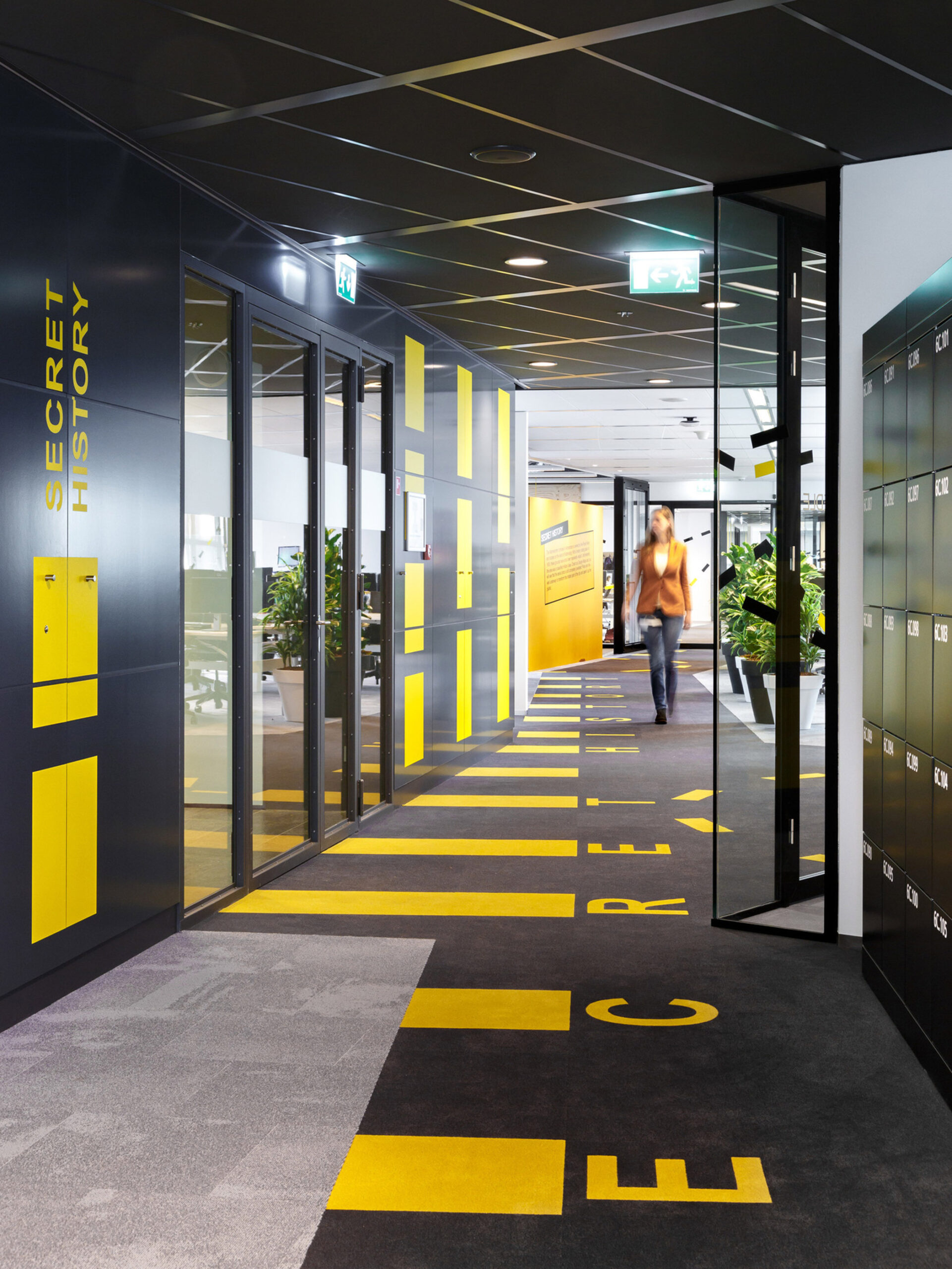 Sleek office corridor with bold yellow graphic elements on a gray floor, accentuating directional flow and zone demarcations, complemented by dynamic lighting and clear signage for a modern, energetic workplace environment.