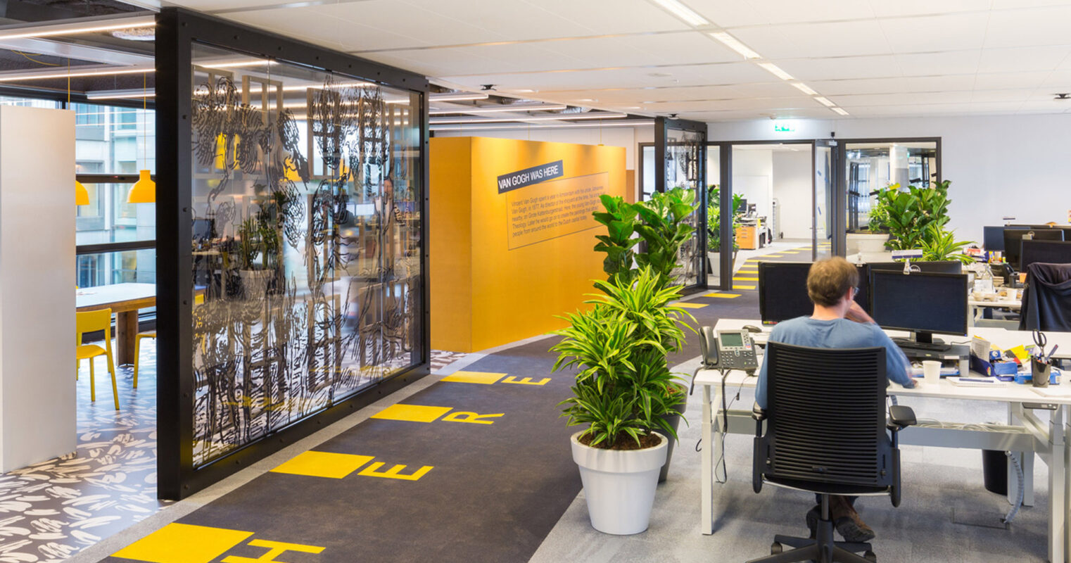 Modern office interior with open-plan layout, featuring a combination of warm wooden tones and vibrant yellow accents. Black-framed glass partitions provide a transparent division, with ergonomic seating and eclectic floor patterns enhancing the dynamic workspace atmosphere.