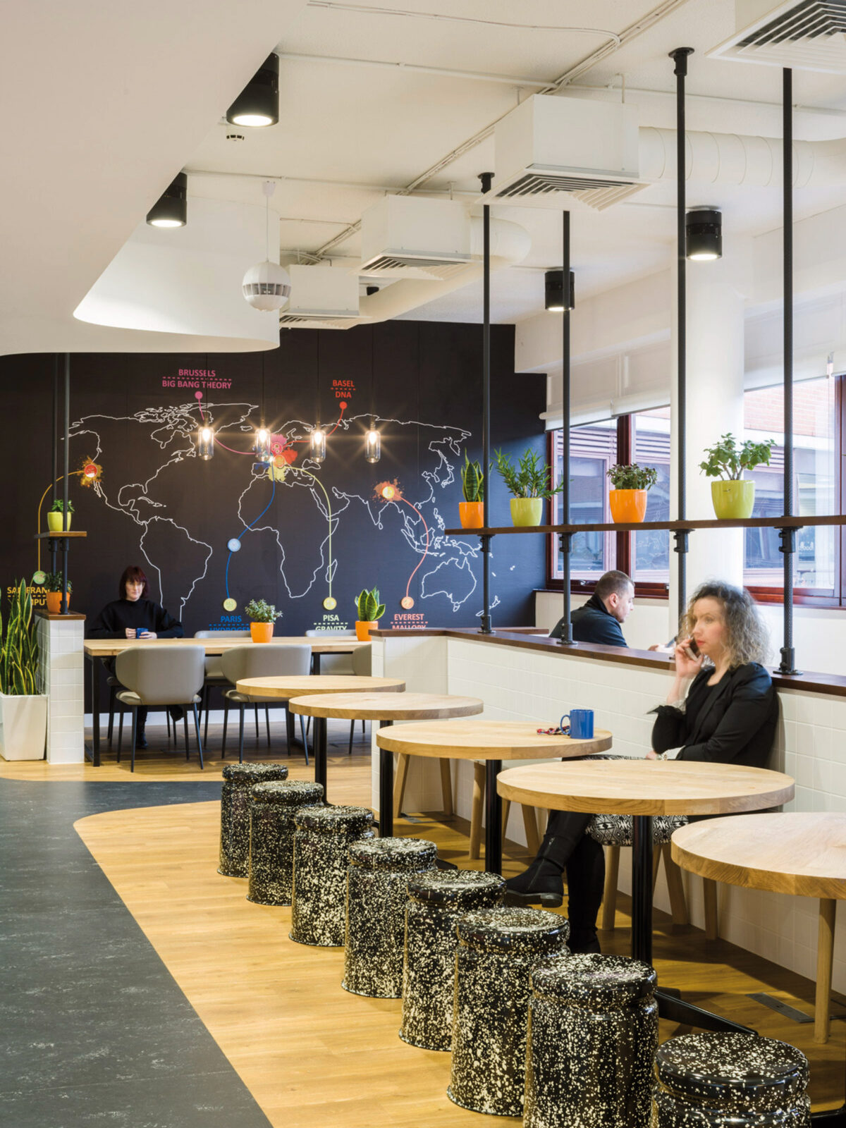 Modern cafeteria space featuring black walls adorned with a vibrant world map mural, eclectic pendant lighting, and a mix of terrazzo stools and wooden tables. Natural light complements the bright accents of decorative planters, merging global inspiration with contemporary design.