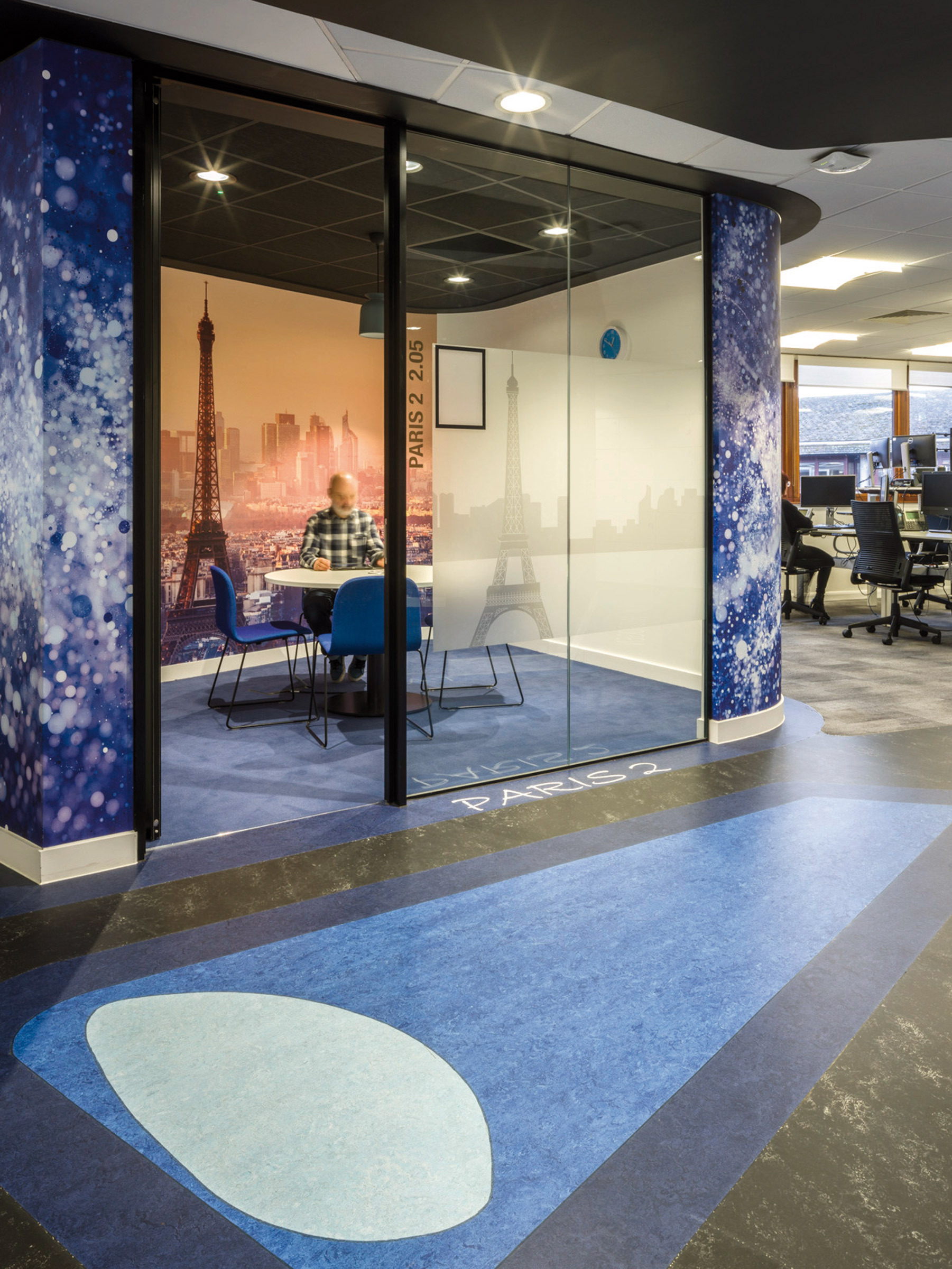 A modern office space featuring floor-to-ceiling glass partitions with an overlaid Paris scene, accented by cosmic and cityscape graphics. The design incorporates a deep blue carpet with a circular pattern, complementing the room's Parisian theme and offering a vibrant, creative work environment.