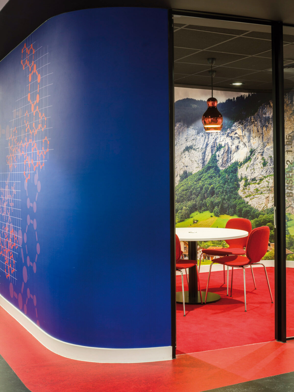 Vibrant interior featuring a curved blue wall with geometric patterns leading to a cozy dining nook with red chairs, a pendant light, and a large scenic photograph of a mountainous landscape.