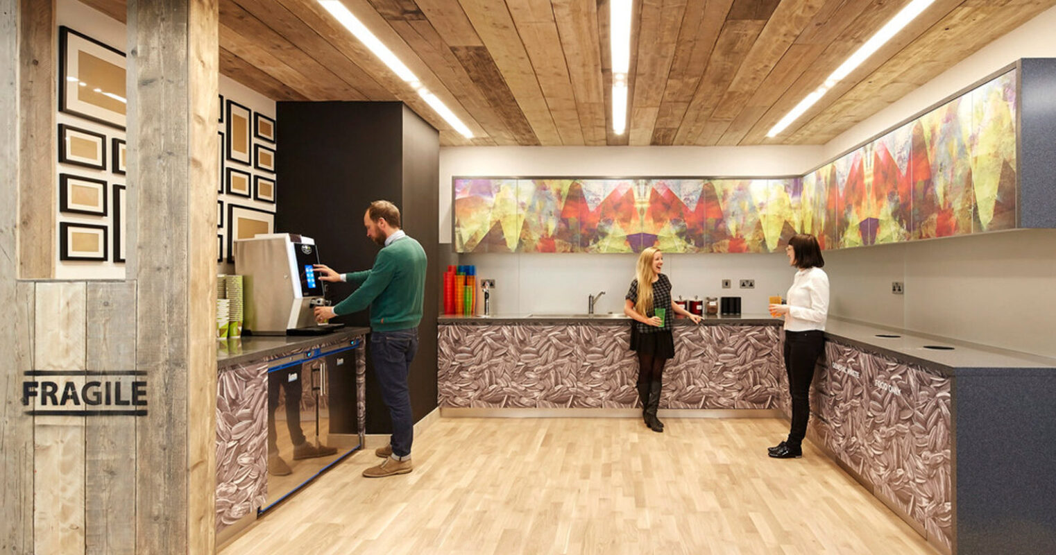 Contemporary office lobby featuring a rustic wood paneled reception desk with abstract textured front panels. Above, a geometric wooden ceiling installation mimics the angular lines of the blue carpet below, complementing the vibrant abstract wall art and creating a dynamic, inviting entrance space.