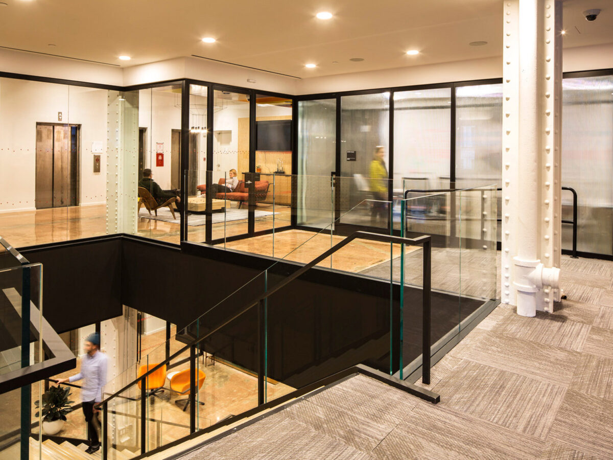 Modern office lobby featuring transparent glass partitions, sleek metal frames, a geometric-patterned carpet, with a neutral color palette accented by warm lighting and wood tones. A person is in motion, walking through the space, enhancing the image's dynamic feel.