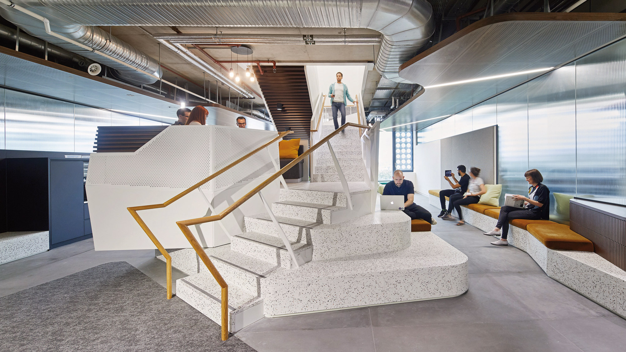 Modern office interior featuring a central floating staircase with white steps and gold accents. The surrounding open-concept space includes a lounge area with mustard yellow seating, exposed ductwork overhead, and people engaged in casual conversation, embodying a contemporary work environment.
