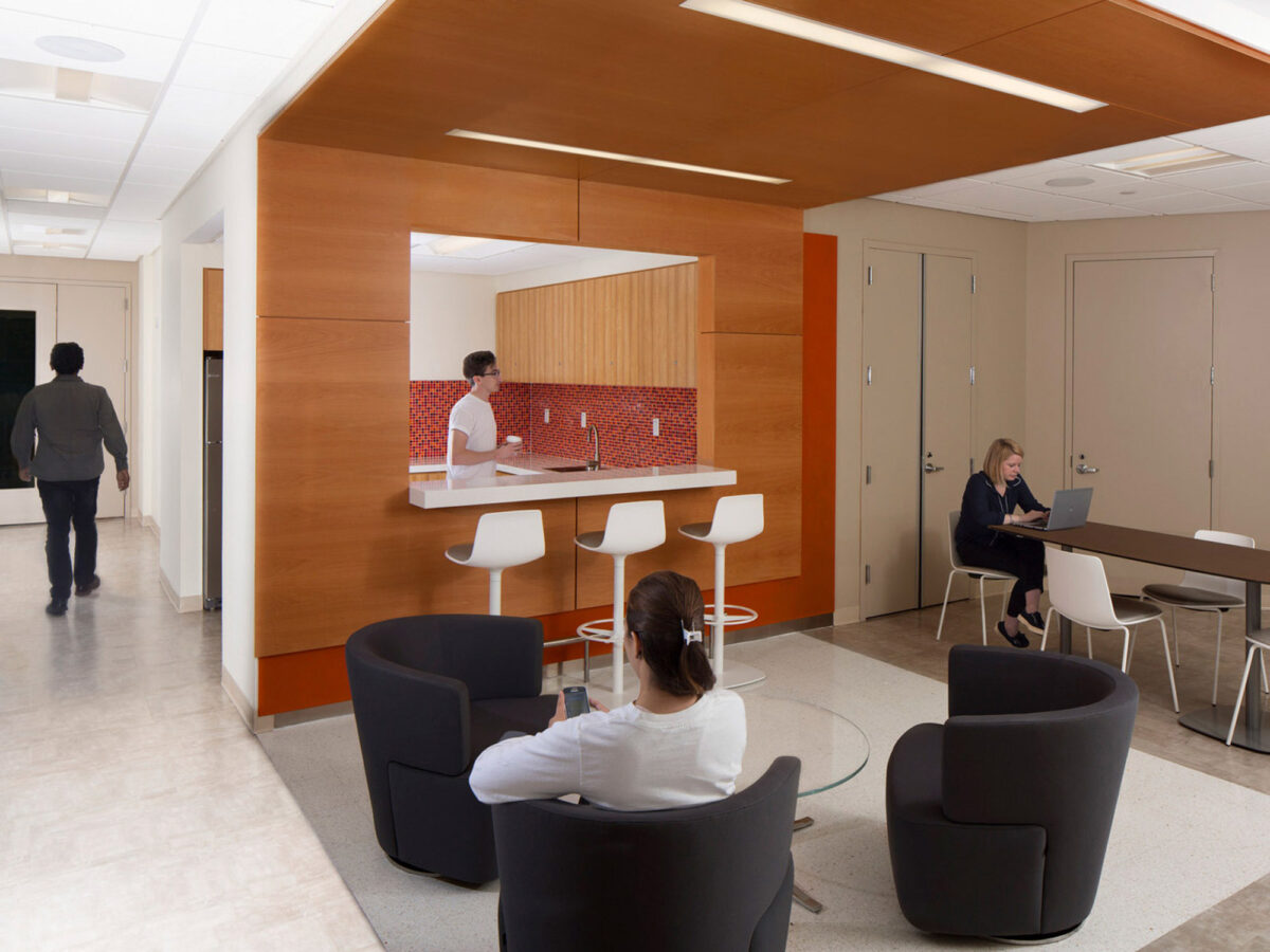 Modern office lounge with white walls and warm wood paneling. Central seating area features four black leather chairs around a glass coffee table, with a woman working on a laptop. A kitchenette with red mosaic backsplash and bar stools is visible in the background.