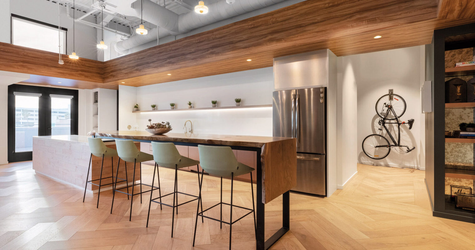 Modern kitchen with a blend of natural wood tones and sleek white cabinetry, featuring an island with bar seating, stainless steel appliances, and herringbone wood flooring. Exposed ductwork adds an industrial touch above, complementing the warm, contemporary aesthetic.