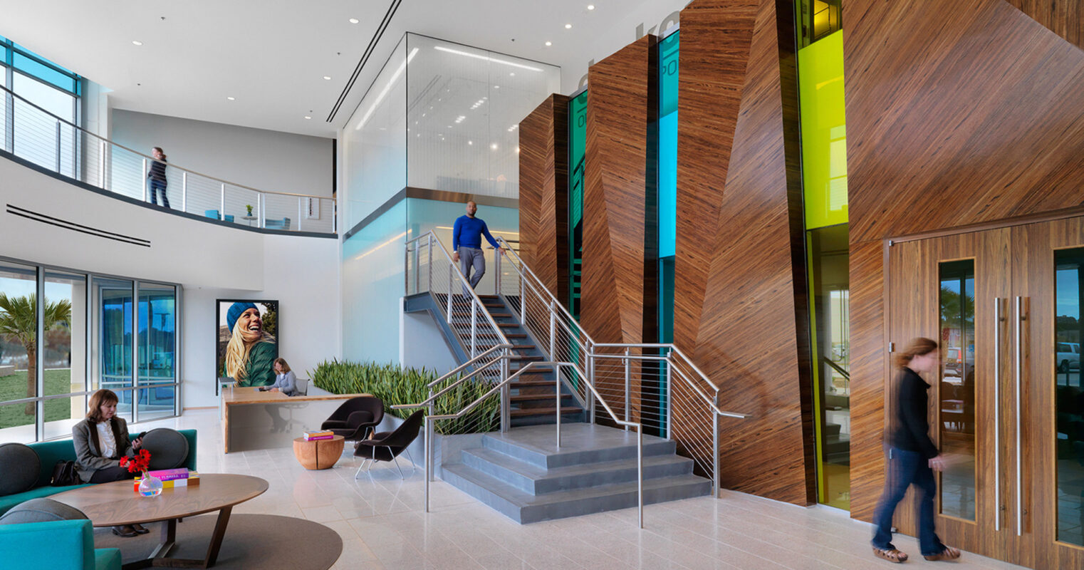 Modern office lobby with high ceilings and a spacious layout. A striking wood feature wall adds warmth, complemented by colorful glass panels. Contemporary furniture provides casual seating, and a metal staircase offers access to the second level, enhancing the airy and dynamic design aesthetic.