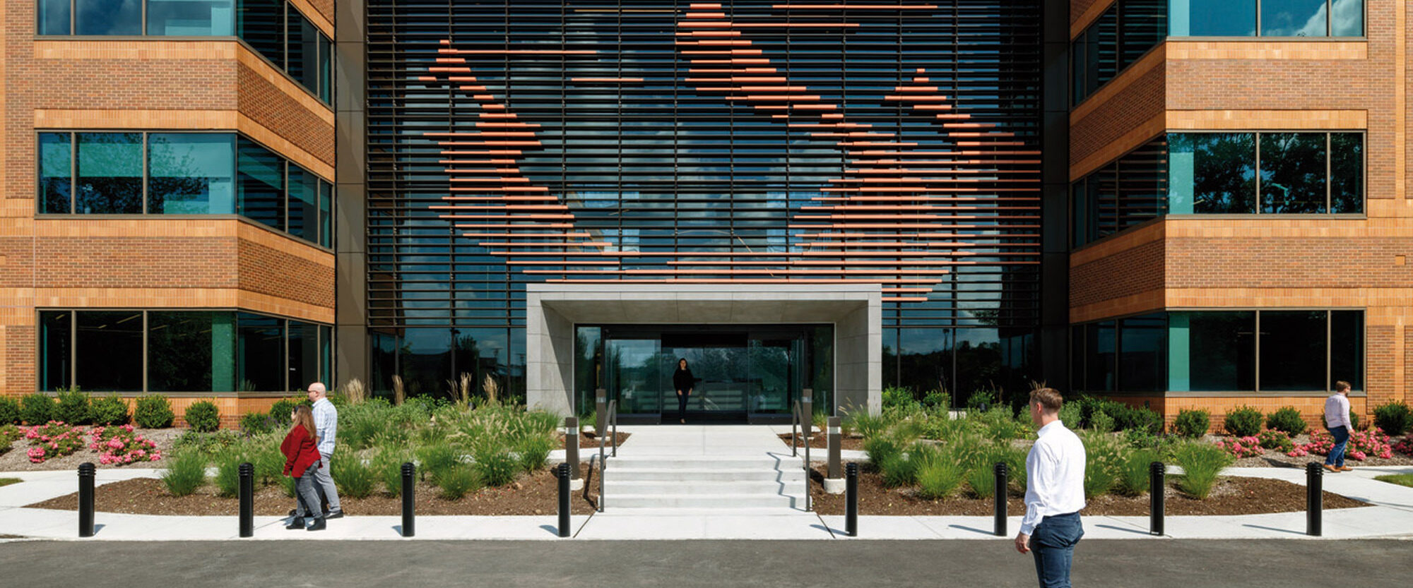 Modern corporate building facade with horizontal copper cladding patterns, flanked by landscaped greenery, with pedestrians approaching a glass entrance.