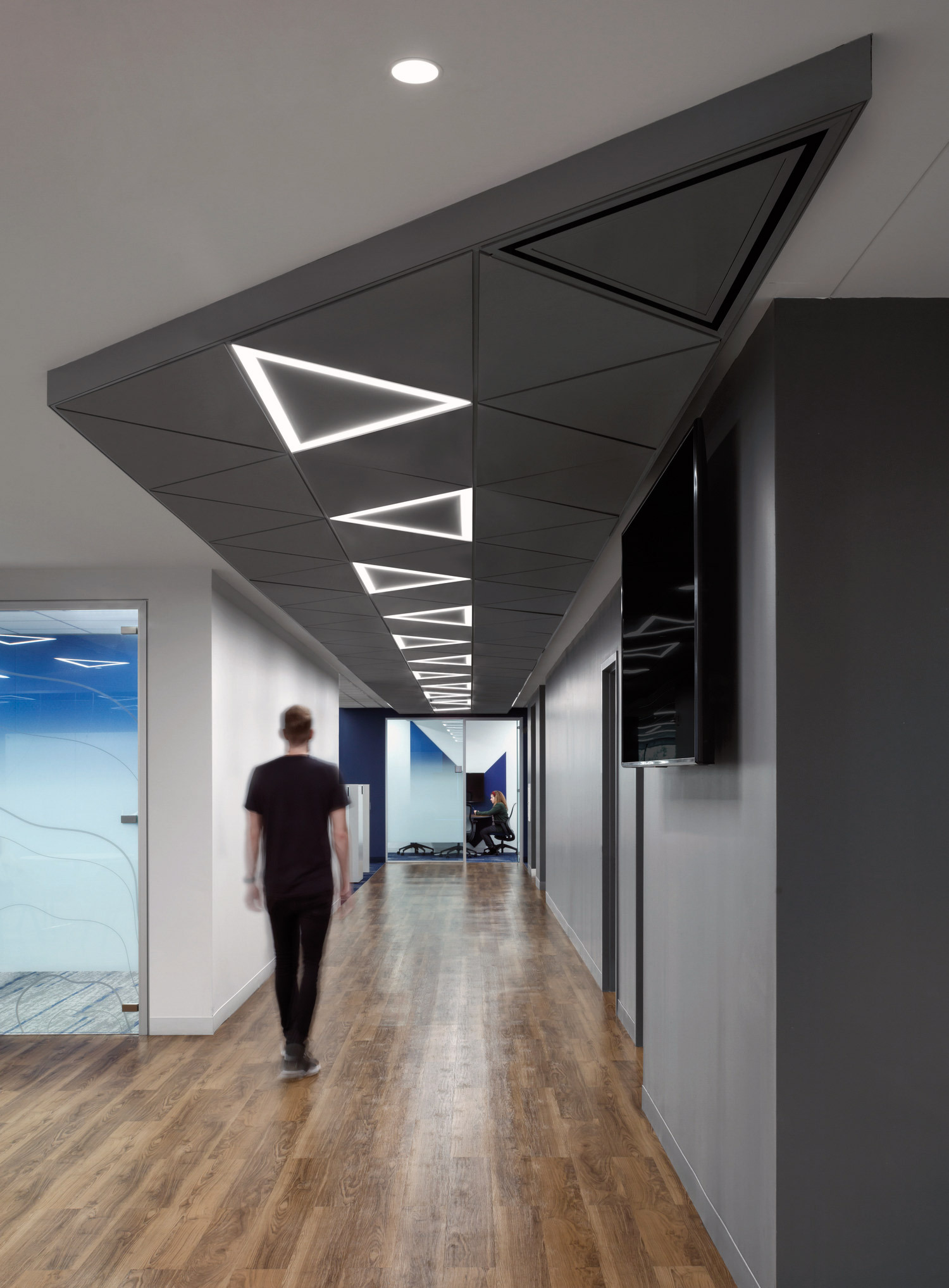 Modern office corridor featuring geometric ceiling with integrated triangular lighting, sleek black panels, polished wooden flooring, and clear glass doors revealing a hint of the vibrant workspace within.