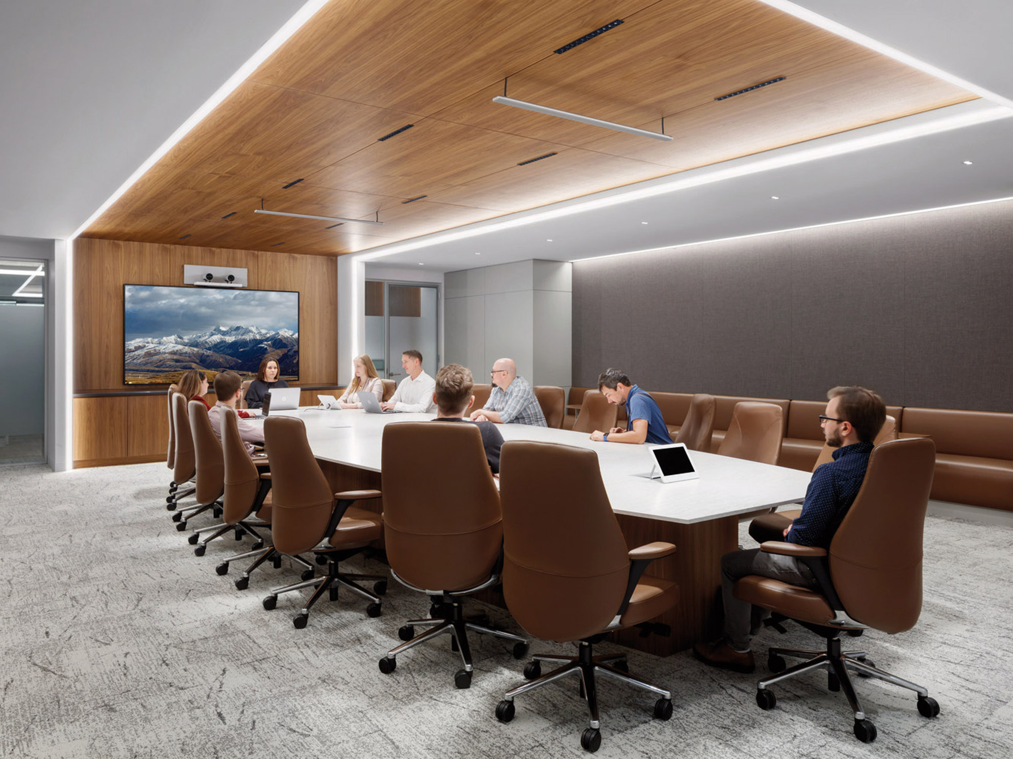 Modern conference room with a sleek, elongated table, leather chairs, and a recessed ceiling with warm wood accents. Ambient lighting complements the neutral color palette, while a large digital screen adorns the front wall, enhancing the collaborative workspace.