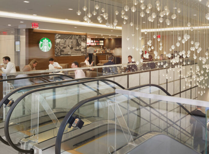 Modern commercial interior featuring an expansive, illuminated ceiling installation with cascading spherical lights over a dual staircase. Reflective surfaces and minimalist furniture provide a sleek, contemporary ambiance, complemented by a coffee shop counter on the upper level.