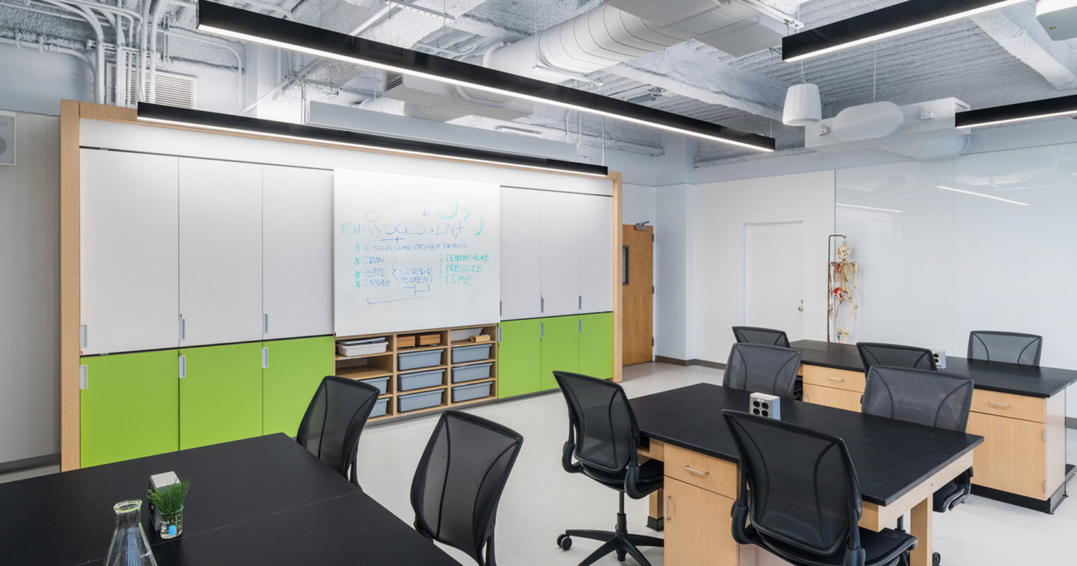Modern office meeting area featuring exposed ceiling infrastructure, whiteboard cabinetry, black rolling chairs, dark tables, and vibrant green accents. Natural light enhances the space's airy feel and complements the minimalist design approach.