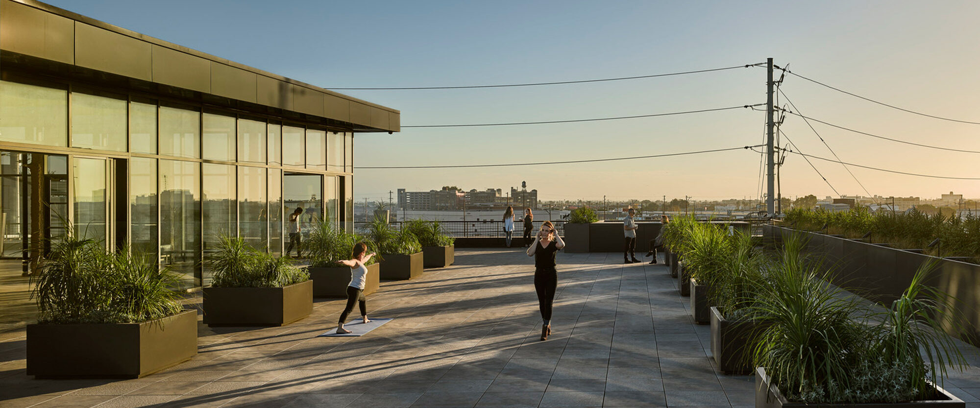 Rooftop terrace with expansive skyline views, featuring large, square planters and minimalist, linear landscaping. Floor-to-ceiling glass facade on the adjoining building enhances the seamless indoor-outdoor aesthetic. Natural light bathes the space, highlighting the clean, architectural lines.