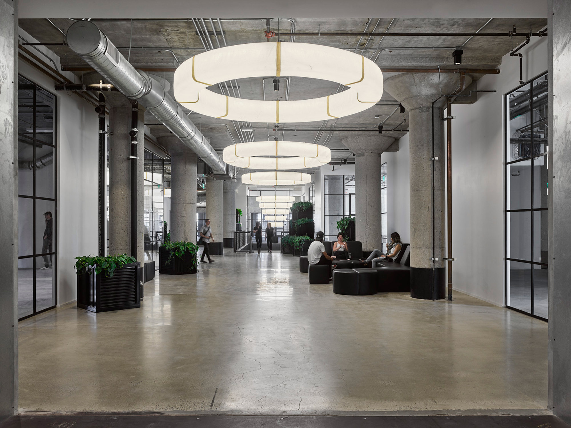 Modern industrial office space with exposed ductwork and concrete floors, enhanced by circular overhead lighting fixtures. Greenspaces with potted plants add vitality, complementing the muted color palette and expansive, open-plan layout promoting collaboration.