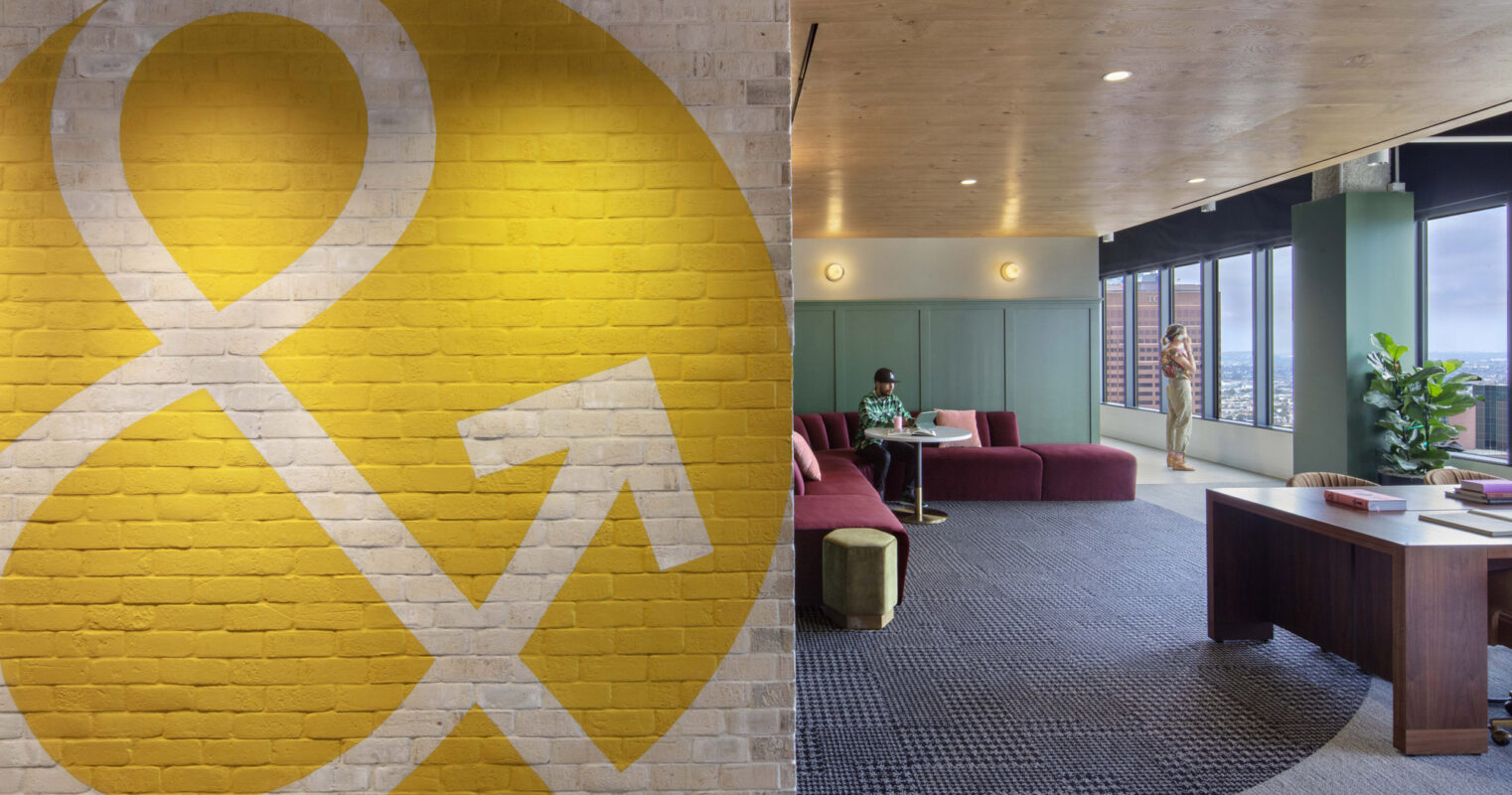 A vibrant yellow accent wall featuring a bold ampersand graphic anchors a modern office lounge. The space balances natural materials with pops of color, offering a mix of private seating areas and a communal workspace with city views.