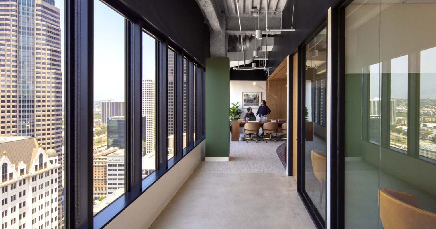 Modern office space boasting floor-to-ceiling windows that invite abundant natural light, framing city views with an open-concept work area and exposed ceiling ductwork, highlighting an industrial design aesthetic.