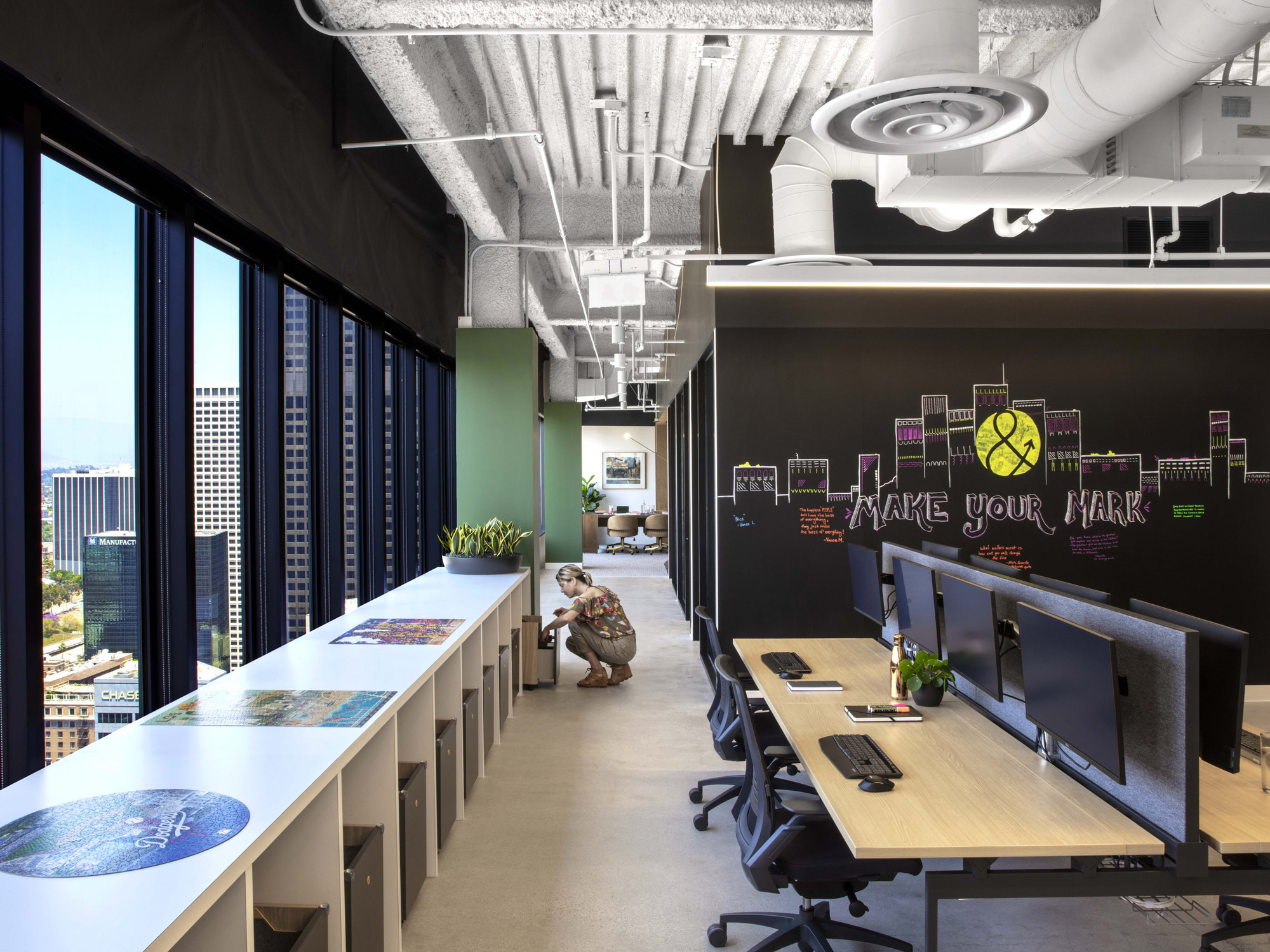 Modern office space with expansive city view, featuring exposed ceiling ductwork, floor-to-ceiling windows, and a vibrant, graffiti-style mural with the text "Make Your Mark." Individual workstations are complemented by colorful area rugs and open collaborative areas.