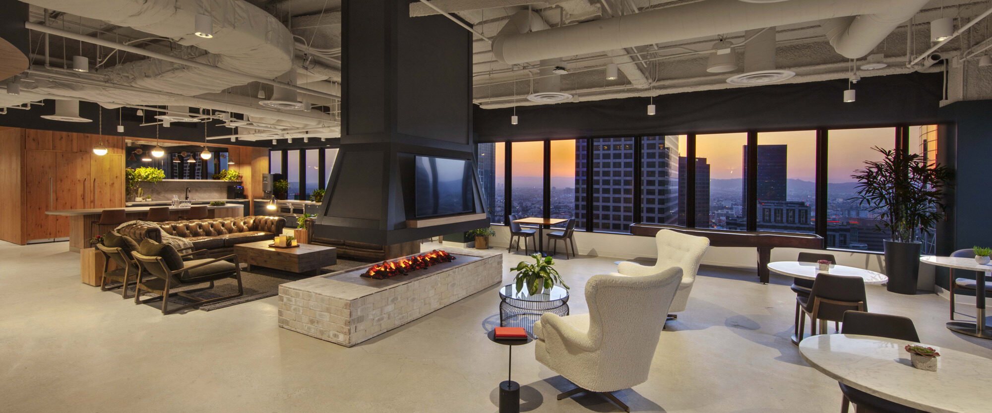 Open-plan office lounge featuring eclectic furnishings, with a warm fireplace, exposed piping on the ceiling, and floor-to-ceiling windows showcasing a cityscape during twilight.