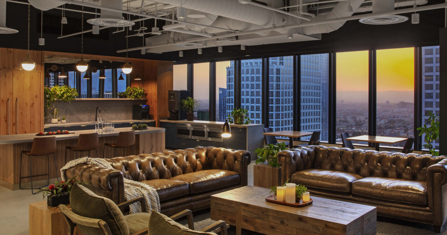 Modern urban loft featuring exposed ceiling ducts and concrete beams, with a central tufted leather Chesterfield sofa, complemented by vintage wooden armchairs and industrial-style lighting. Floor-to-ceiling windows offer a panoramic city skyline view at dusk.
