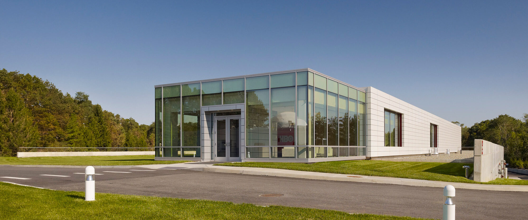 Modern glass-fronted building with clean lines and minimalist design, set against a clear blue sky and situated in a landscaped environment with green lawns and sparse trees. The structure features a flat roof and expansive windows allowing ample natural light.
