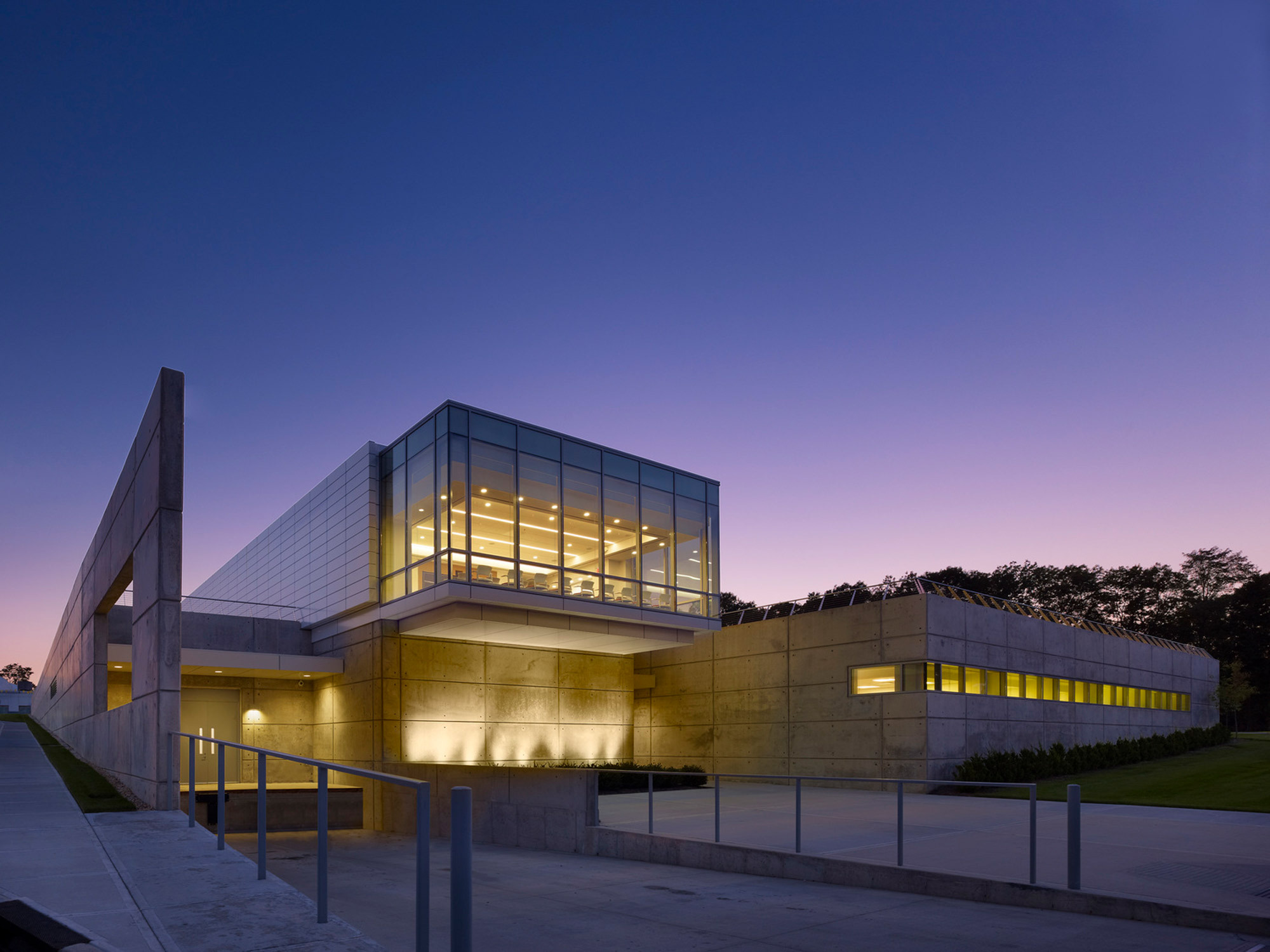 Modern architectural building at dusk with illuminated interiors visible through large windows. The structure features clean lines, cantilevered upper level, and subtle accent lighting highlighting its geometric form against a gradient twilight sky.