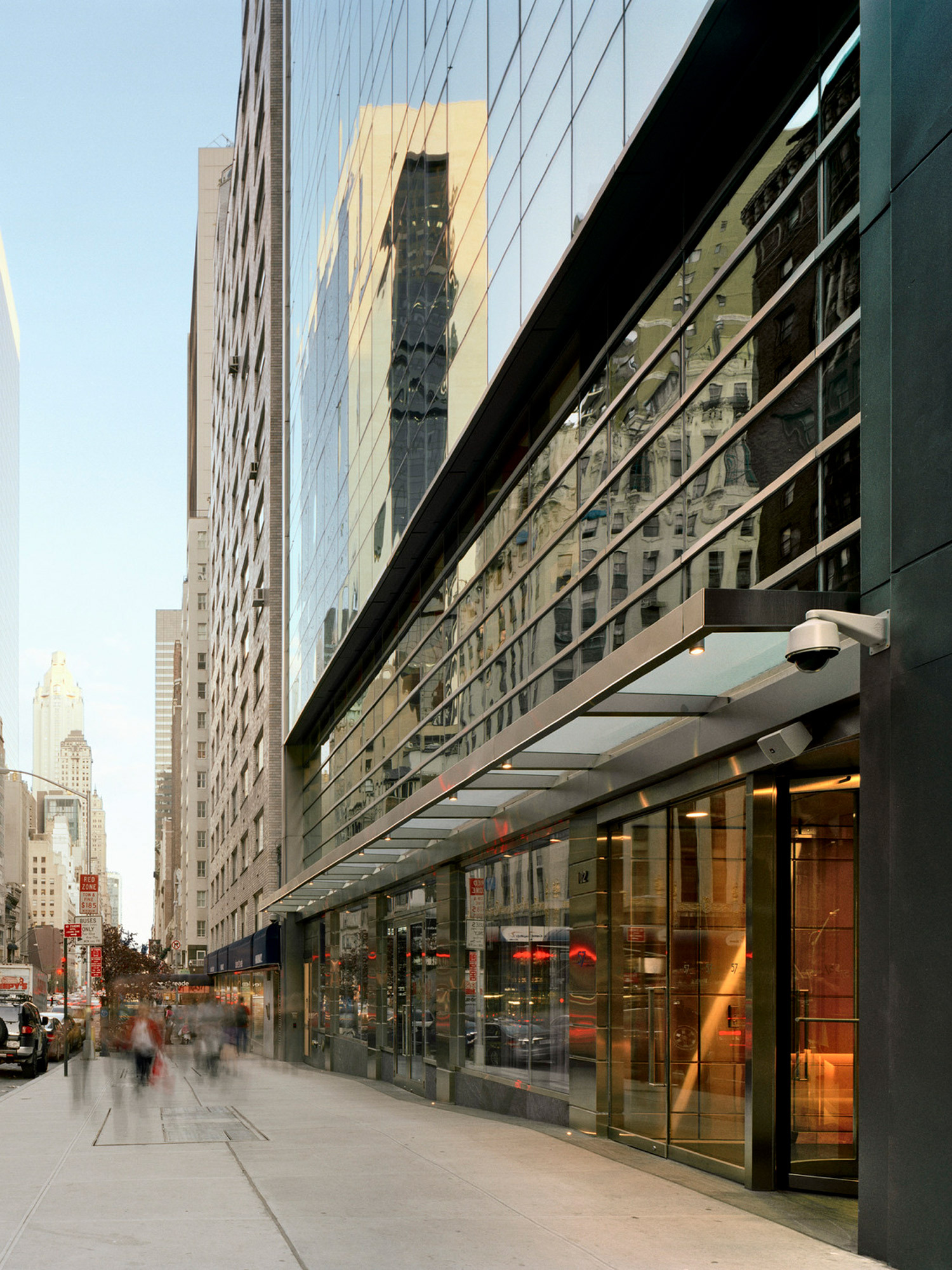 Exterior view of a modern building featuring reflective glass facade and asymmetrical windows, with pedestrians blurred in motion on a vibrant city street. The entrance boasts a sleek, overhanging canopy and recessed lighting, highlighting contemporary urban architecture.