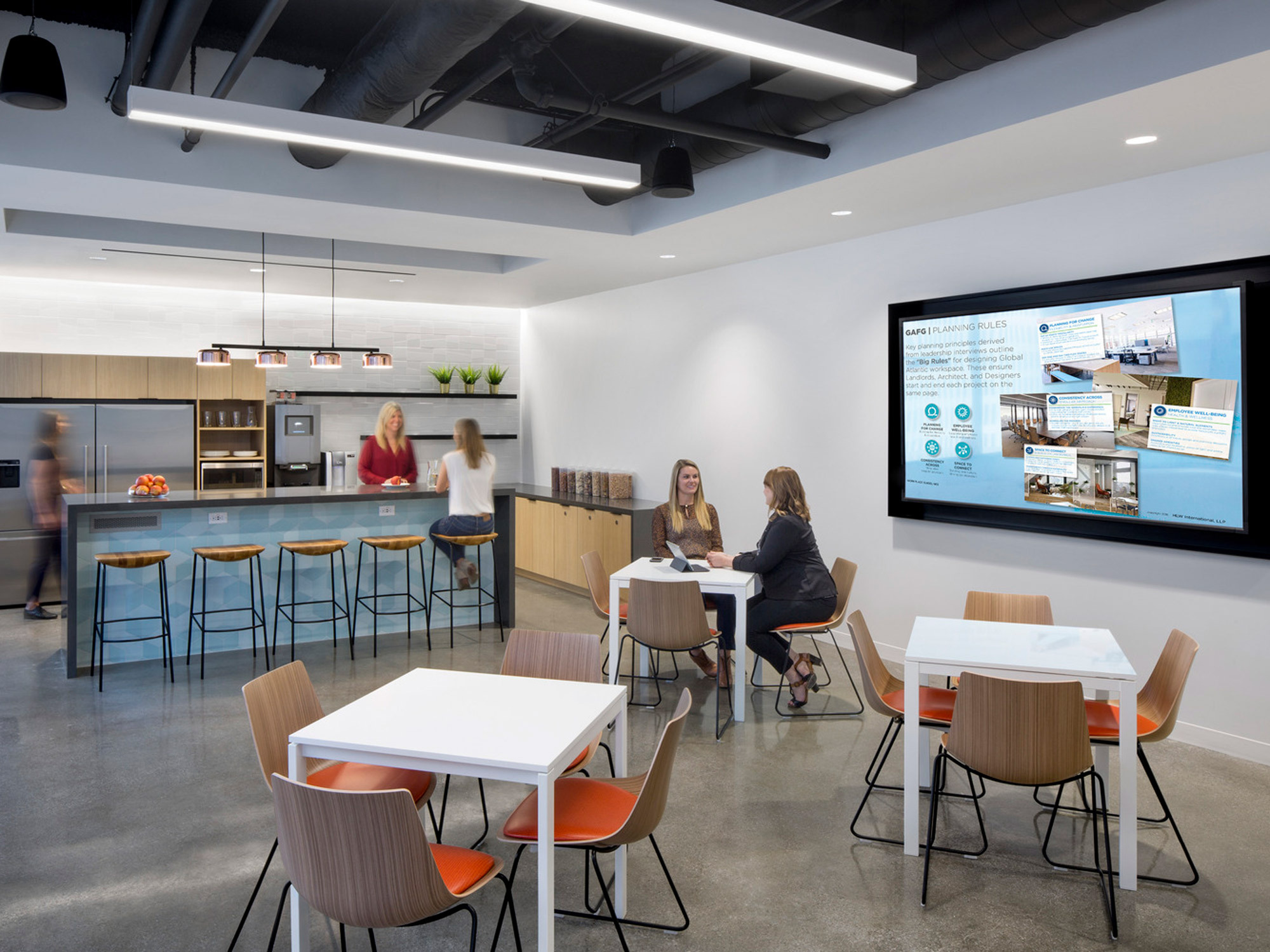 Modern office break room featuring sleek, two-tone kitchen cabinetry and bar-seating area with orange stools. An interactive digital display presents information, while employees engage in casual conversation around tables with white chairs. The space combines functionality with a stimulating color palette for collaborative work.
