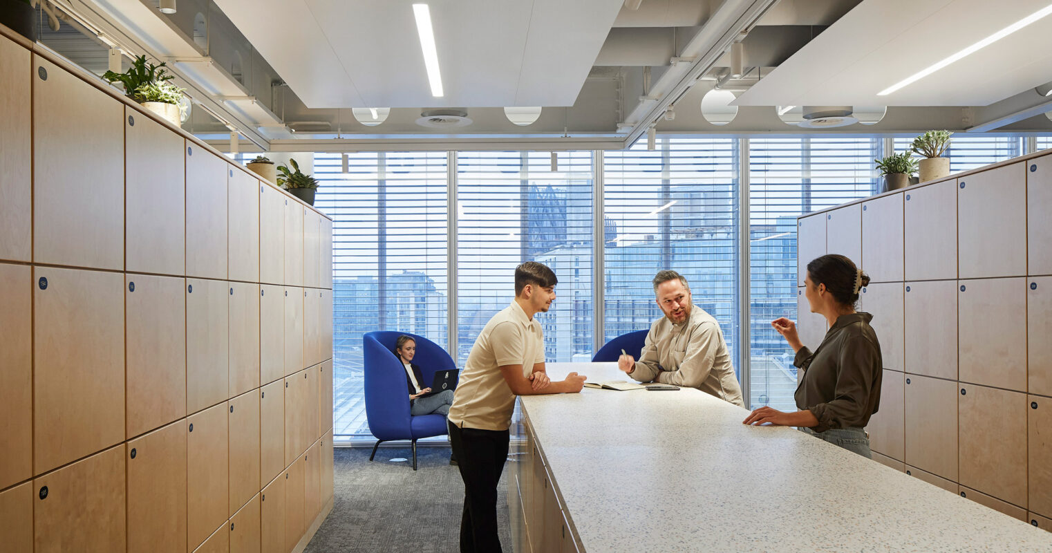 Modern office space showcasing neutral wood lockers, terrazzo-style countertops, and recessed lighting. Three professionals engage in a discussion over documents at the central white table, adding a sense of collaboration to the clean, structured environment.