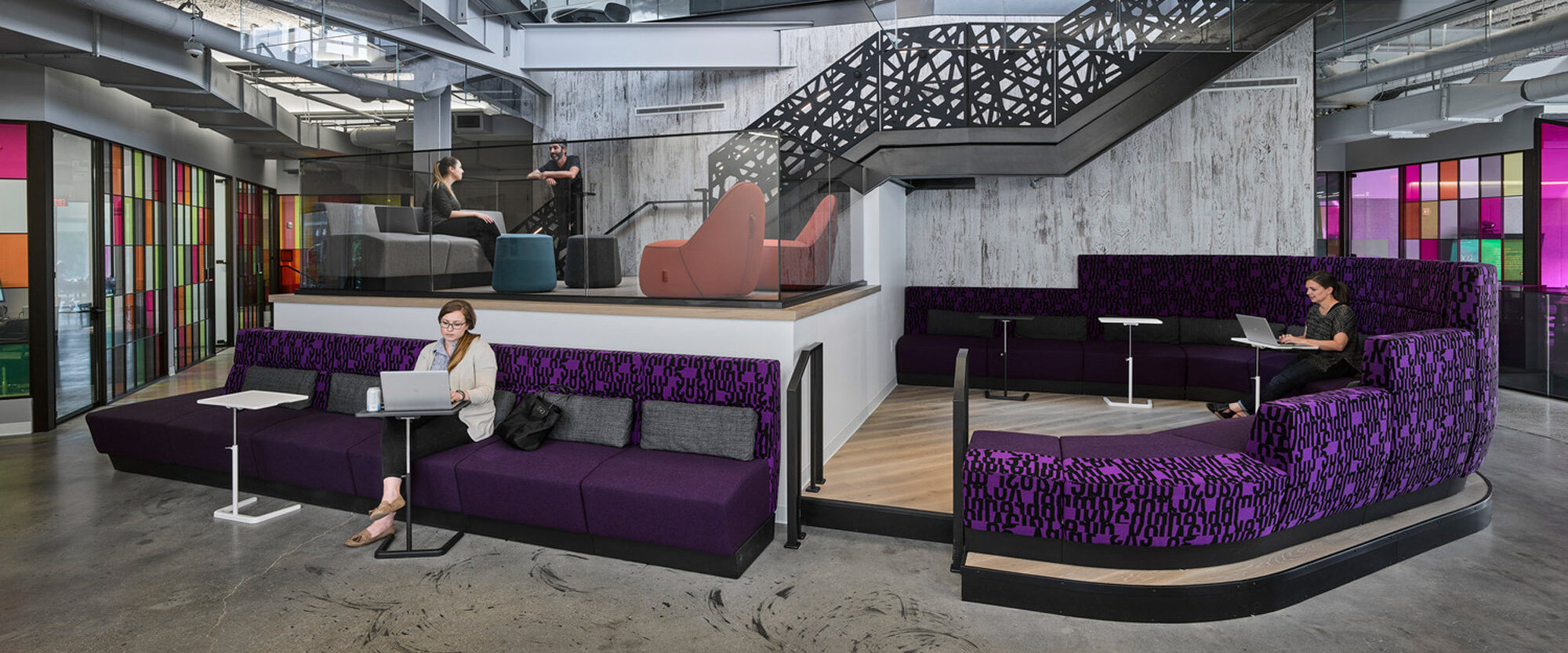 Modern office lobby featuring eclectic furnishings with purple upholstery patterns, a central geometric staircase with metal finishes, and vibrant stained-glass panels contributing to a dynamic and creative workspace atmosphere.