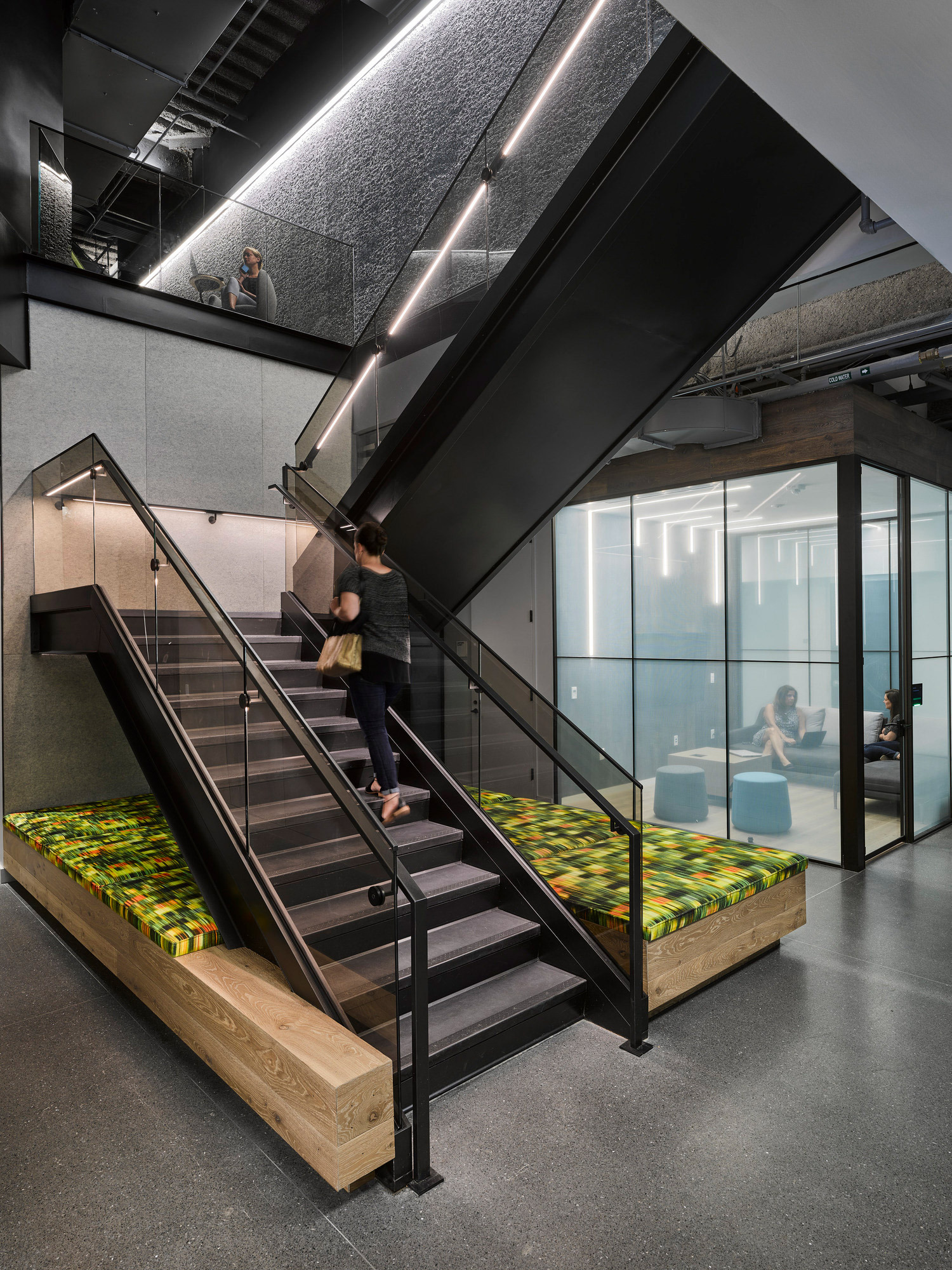 Modern office stairway linking two levels, featuring black metal railings, wood accented steps with vibrant green foliage patterned carpet inlay, set against an industrial concrete and exposed black ceiling backdrop, with glass-walled meeting rooms visible in the background.