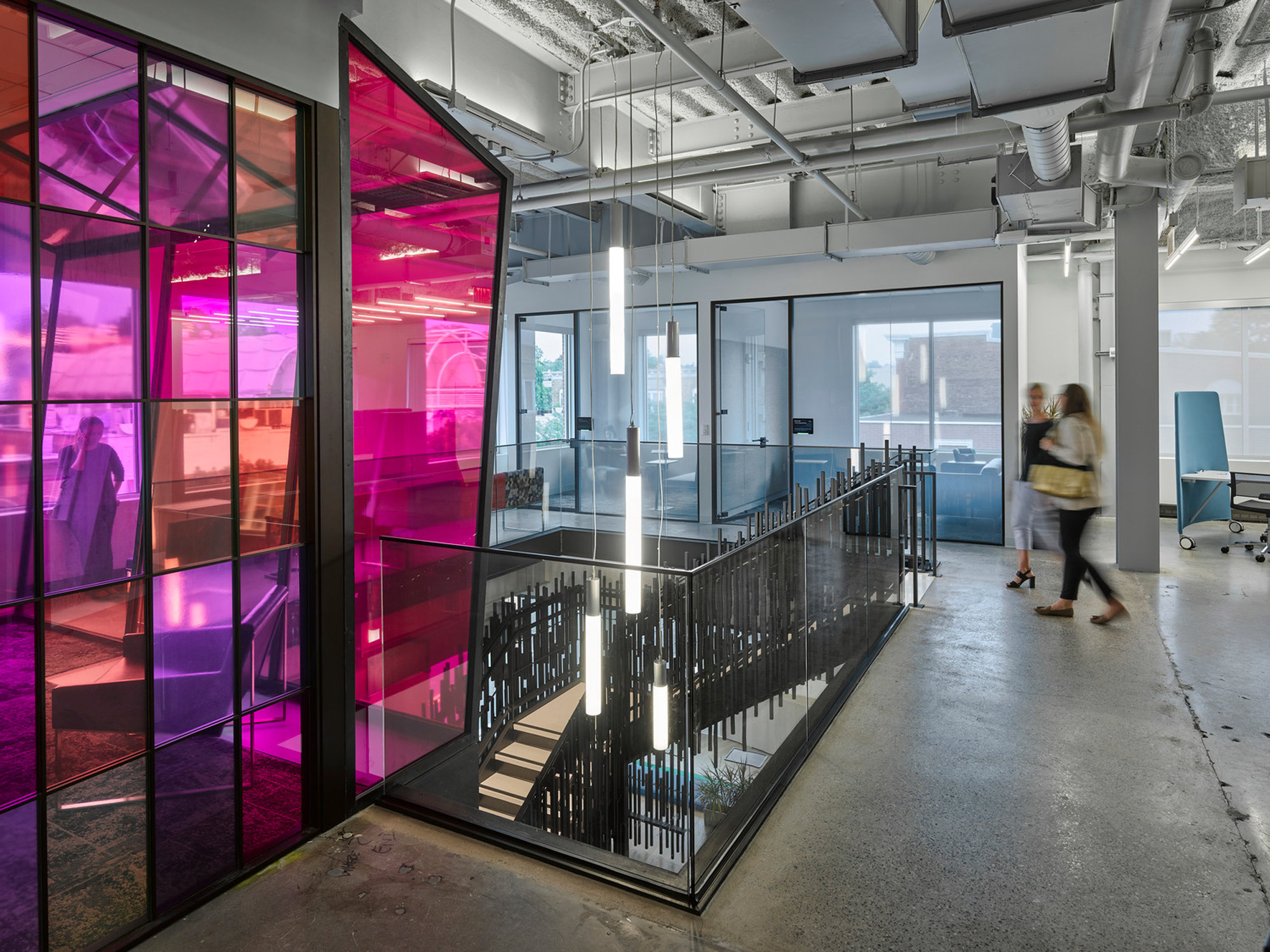Modern office interior featuring translucent pink glass partitions, creating a dynamic contrast with the industrial aesthetic of exposed pipes and ductwork. A sleek black staircase with railing descends into the vibrant space, complemented by the natural light streaming through large windows.