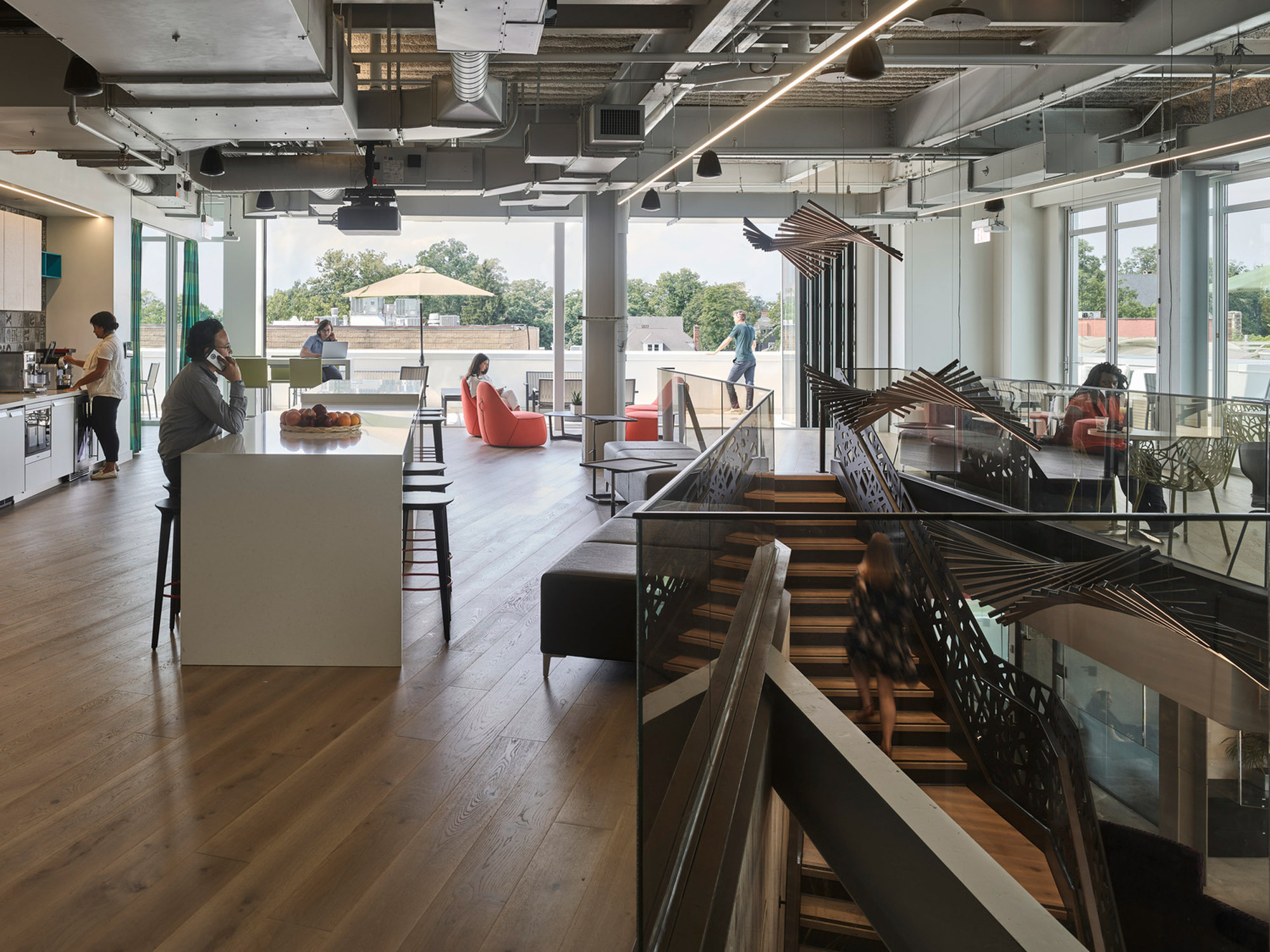 Open-plan office space with modern design, featuring exposed ceiling ductwork, polished concrete flooring, and contemporary furniture. A kitchenette with bar seating takes foreground while employees engage in various activities within natural light-filled surroundings. Unique wooden partition sculptures provide visual interest and spatial division.